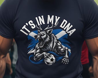May include: Navy blue t-shirt featuring a wolf-like creature with antlers, holding a soccer ball, and the text "IT'S IN MY DNA." The design incorporates the Scottish flag. The shirt is a dark blue color.