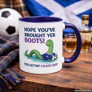 Può includere: Tazza in ceramica bianca con interno e manico viola. Illustrazione di un mostro di Loch Ness verde con maglia da calcio, che tiene un pallone. Testo: "HOPE YOU'VE BROUGHT YER BOOTS!" e "YER GETTIN' CALLED OAN!". Tema scozzese.
