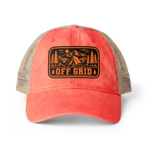 Op de afbeelding: Een koraalkleurige truckerpet met een beige mesh achterkant en een rechthoekige patch op de voorkant. De patch bevat een zwarte en oranje afbeelding van een tent, bergen, bomen en een kampvuur met de tekst OFF GRID in dikke hoofdletters.