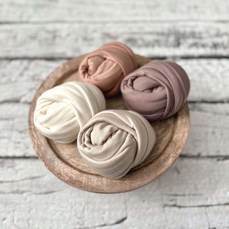 May include: Four swaddles in shades of cream, peach, and brown are arranged on a wooden plate. The swaddles are tightly rolled, creating a textured appearance. The plate has a rustic, worn look, and the background is a white brick wall.