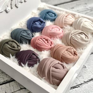 May include: A white wooden tray filled with a fluffy white material, holding several rolled-up fabric wraps in various colors. The wraps include shades of blue, pink, green, and brown, perfect for newborn photography.