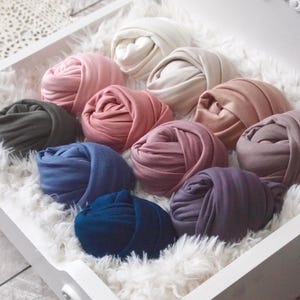 May include: A collection of rolled fabric wraps in various colors, including pink, blue, and beige, arranged in a white tray with a fluffy white lining. The wraps are tightly wound, showcasing their soft texture and diverse hues.