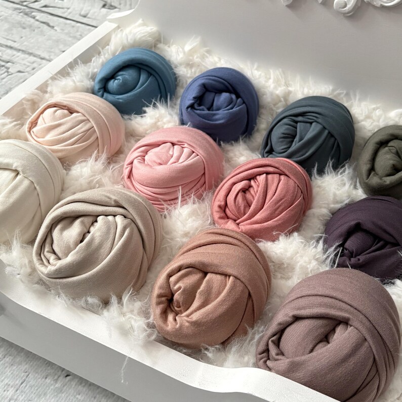 May include: A collection of rolled fabric in various pastel colors, including pink, blue, and beige, arranged in a white tray filled with fluffy white material. The fabric appears soft and is likely used for crafting or baby photography.