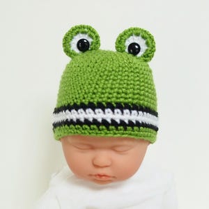 May include: Green crocheted baby hat with black and white stripes and frog ears with black eyes.