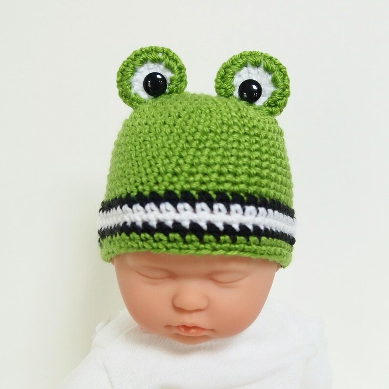 May include: Green crocheted baby hat with black and white stripes and frog ears with black eyes.