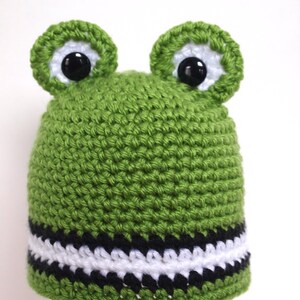 May include: Green crocheted frog hat with black and white stripes around the brim. The hat has large black eyes and green felt ears.