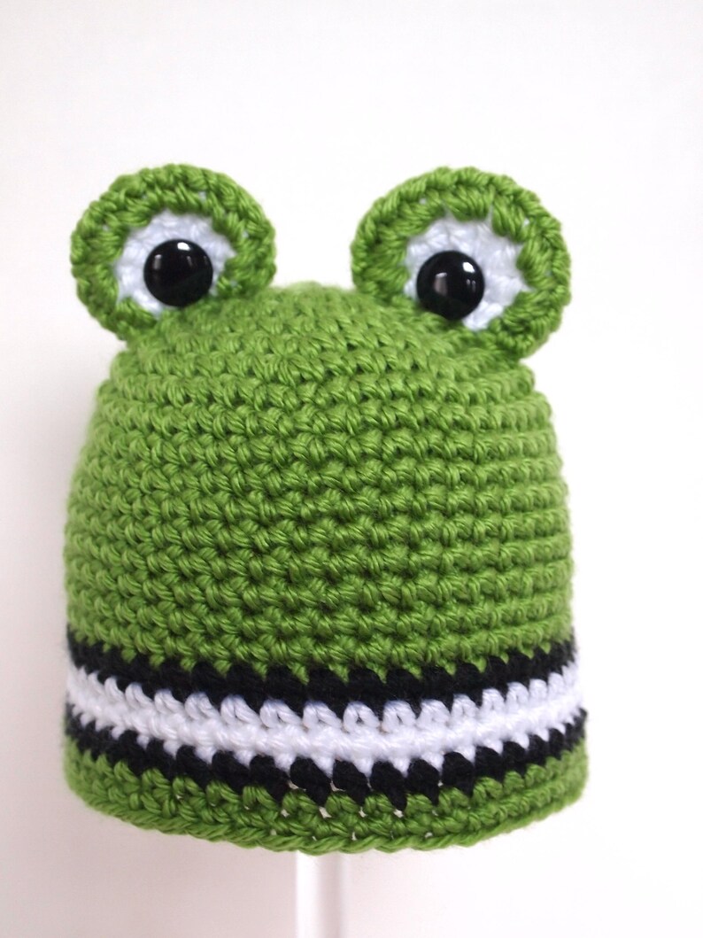 May include: Green crocheted frog hat with black and white stripes around the brim. The hat has large black eyes and green felt ears.