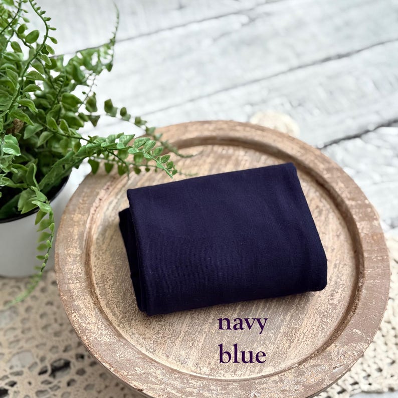 May include: A folded piece of navy blue fabric. The words "navy blue" are printed in a script font on the bottom right corner. The fabric is displayed on a light brown, round wooden tray.