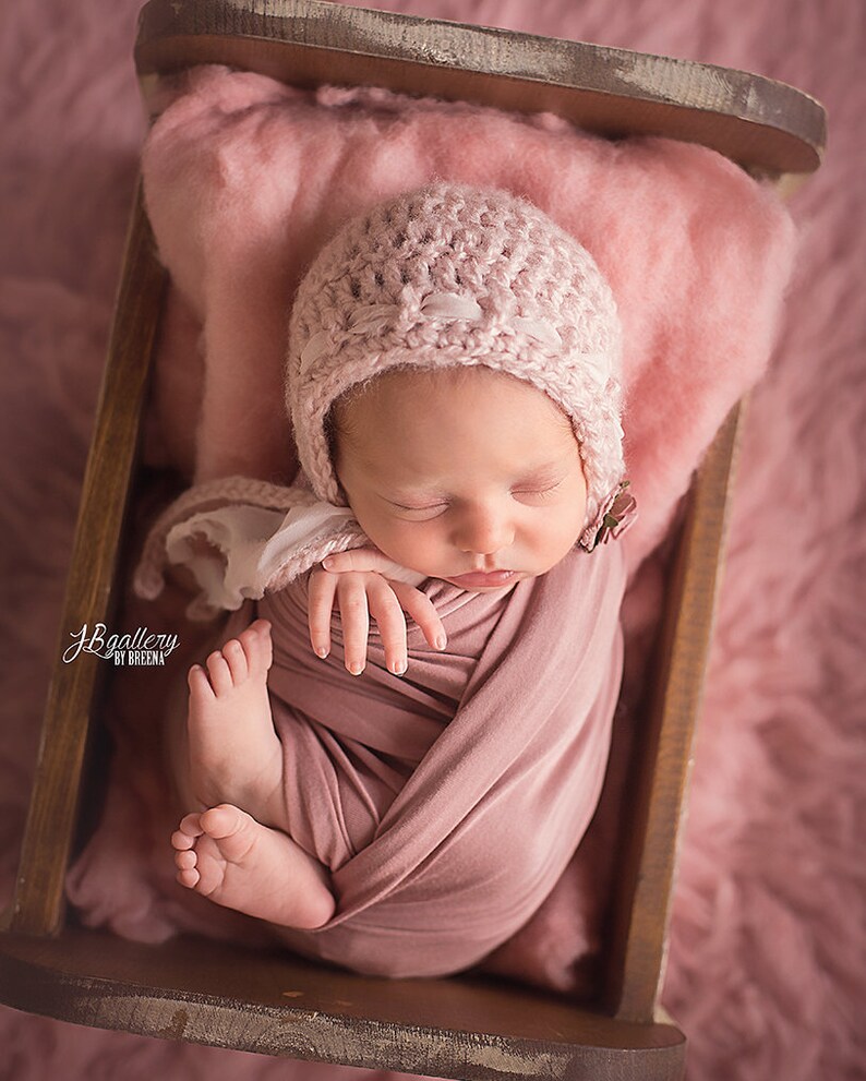 May include: A newborn baby wrapped in a mauve blanket and wearing a pink knitted bonnet, resting in a small wooden bed. The bed is lined with pink fluffy material. The baby is curled up, with tiny feet visible. The image has a soft, warm tone.