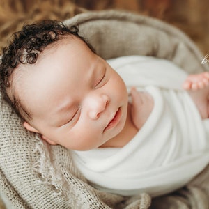 May include: A newborn baby swaddled in a white blanket, nestled in a soft, gray basket. The baby has dark, curly hair and is peacefully sleeping. The background is a blurred, neutral tone.
