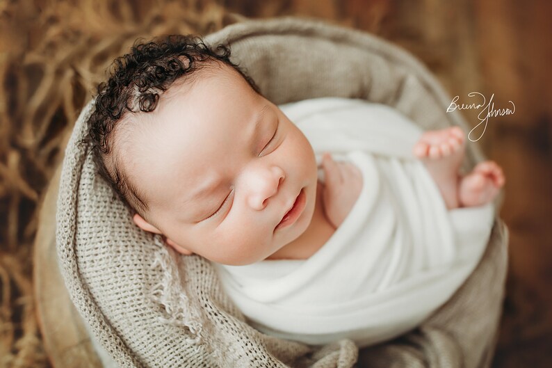 May include: A newborn baby swaddled in a white blanket, nestled in a soft, gray basket. The baby has dark, curly hair and is peacefully sleeping. The background is a blurred, neutral tone.