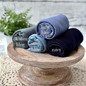 May include: Four rolled fabrics in shades of blue: dk slate blue, steel blue, light blue, and navy. The fabrics are displayed on a rustic, wooden, tiered stand. The names of the colors are printed on the fabric.