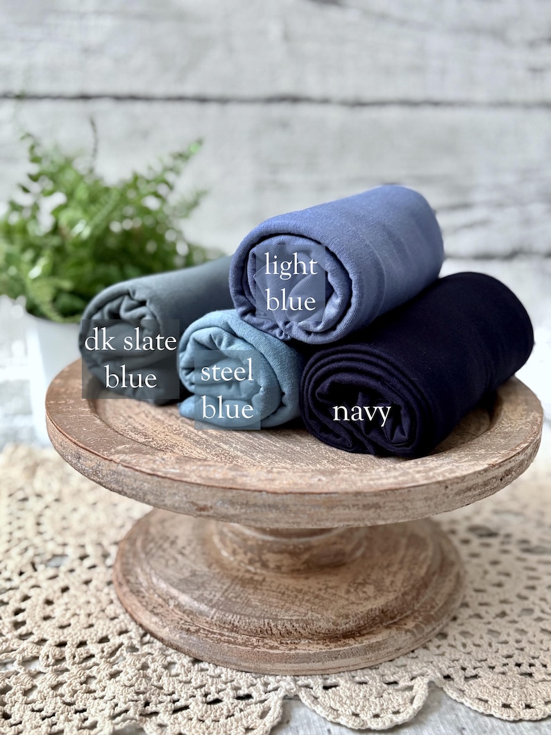 May include: Four rolled fabrics in shades of blue: dk slate blue, steel blue, light blue, and navy. The fabrics are displayed on a rustic, wooden, tiered stand. The names of the colors are printed on the fabric.