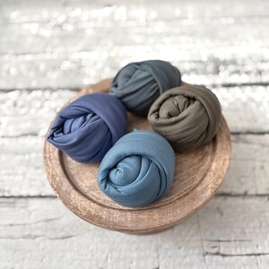 May include: Four rolled fabric wraps in shades of blue and olive green, arranged on a round, wooden tray. The wraps are tightly wound, showcasing their soft texture. The tray has a rustic, weathered appearance, adding a touch of vintage charm.