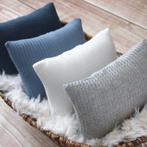 May include: Four rectangular throw pillows in a woven waffle pattern, in shades of navy, blue, white, and gray, rest in a woven basket lined with white faux fur. The pillows are arranged in a row, showcasing their different colors and textures.