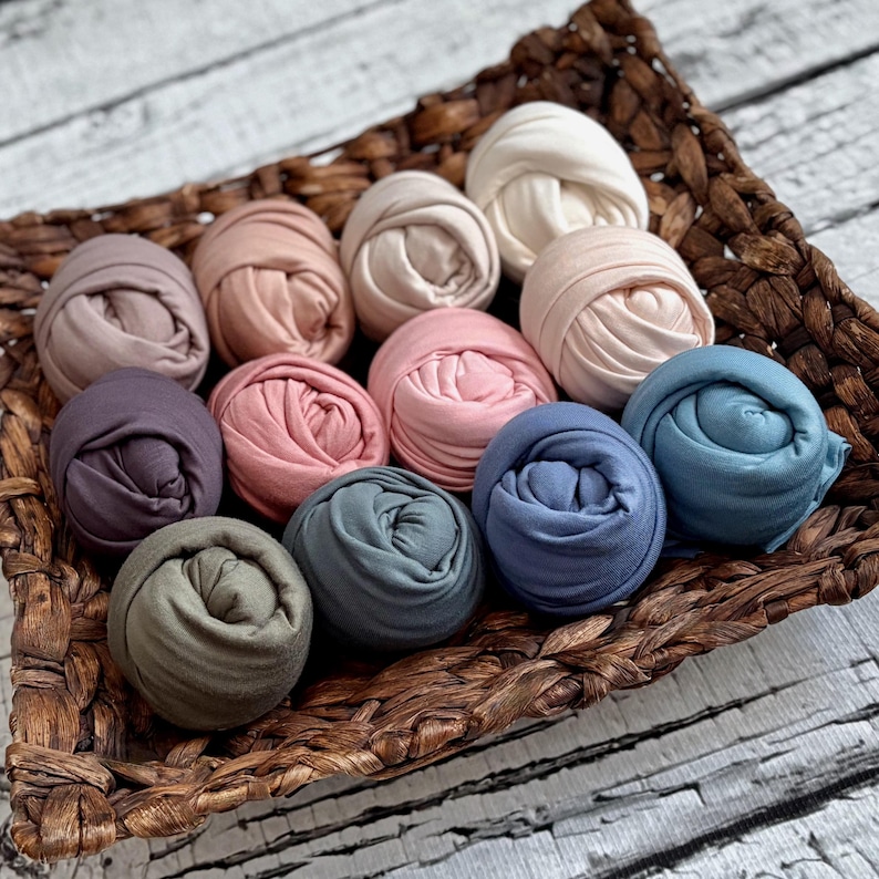 May include: A woven basket holds a collection of rolled fabric in various pastel colors. The assortment includes shades of pink, blue, beige, and purple. The fabric is tightly rolled, creating a textured appearance. The basket sits on a white wooden surface.