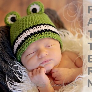 May include: A green crocheted frog hat with black and white stripes and black eyes. The hat is on a baby's head.
