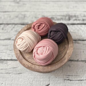 May include: A wooden tray holds five swaddled baby wraps in shades of pink, mauve, and brown. The wraps are tightly rolled and arranged in a circular pattern. The tray has a rustic, weathered appearance.