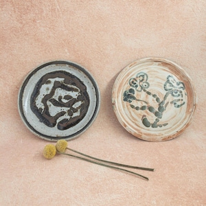 May include: Two handmade ceramic plates with unique designs. One plate is gray with a dark brown abstract pattern, and the other is tan with a blue and brown floral design. Yellow dried flowers are in the foreground.