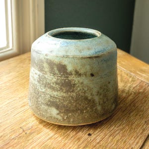 Könnte beinhalten: Eine handgefertigte Keramikvase mit zylindrischer Form und breiter Öffnung. Die Vase weist eine Mischung aus Erdtönen auf, darunter Braun, Grün und Blau, mit einer strukturierten Oberfläche. Die Vase ist etwa 15 cm hoch.
