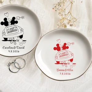 May include: Two white ceramic plates with gold rims. One plate features a black illustration of Mickey and Minnie Mouse in a car with "Just Married" text and the names "Caroline & David" and a date. The other plate has a red illustration with the names "Emma & Alex" and a date.