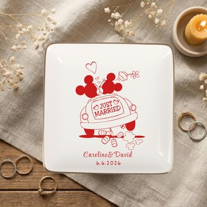 May include: A square, white ceramic ring dish with a gold rim. The dish features a red illustration of a car with Mickey and Minnie Mouse on top, and the words "Just Married." The names "Caroline & David" and the date "6.6.2026" are also printed in red.