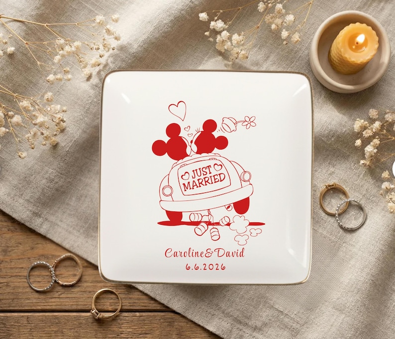 May include: A square, white ceramic ring dish with a gold rim. The dish features a red illustration of a car with Mickey and Minnie Mouse on top, and the words "Just Married." The names "Caroline & David" and the date "6.6.2026" are also printed in red.
