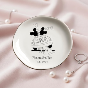 May include: A white ceramic ring dish with a gold rim, featuring a black and white illustration of Mickey and Minnie Mouse in a car with the words "Just Married." The dish is personalized with names and a date.