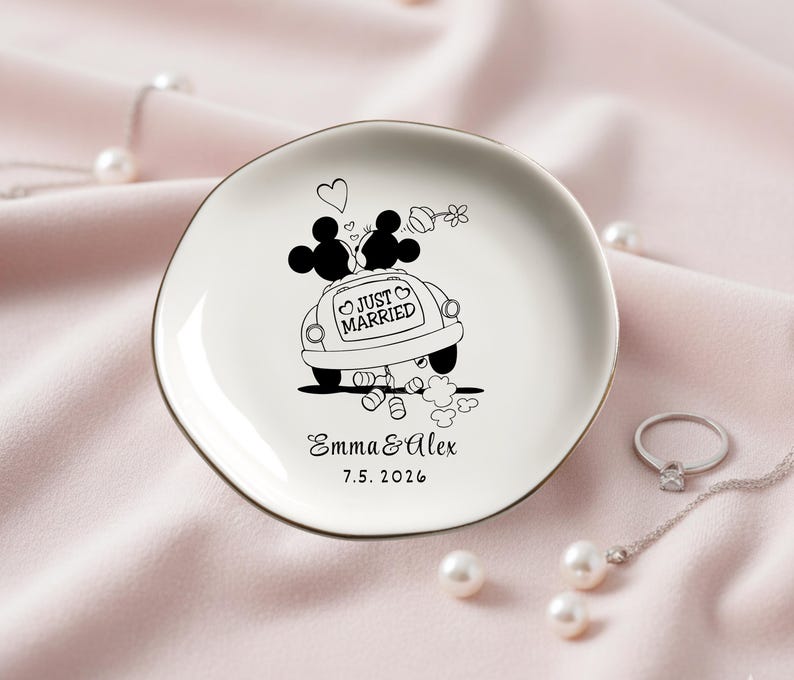 May include: A white ceramic ring dish with a gold rim, featuring a black and white illustration of Mickey and Minnie Mouse in a car with the words "Just Married." The dish is personalized with names and a date.