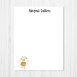May include: A white rectangular sheet of stationery featuring the name Abigail Sutton printed in black script at the top center and a small illustration of a yellow and black striped bee wearing a pink flower crown at the bottom left corner.