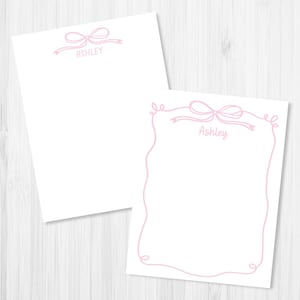 May include: Two white notepads with a pink bow design and the name "Ashley" printed in a delicate script. One notepad has a bow at the top, while the other has a bow and a decorative border. The notepads are on a light wood-grain surface.