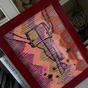 Könnte beinhalten: Ein gerahmtes Kunstwerk mit rotem Rahmen. Das Kunstwerk zeigt ein farbenfrohes abstraktes Design mit rosa, orangefarbenen und gelben Streifen. Schwarze Linien und kleine Blumendetails sind ebenfalls vorhanden und erzeugen eine einzigartige visuelle Textur.