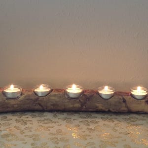 Puede incluir: Un portavelas rústico de madera con cinco velas de té encendidas. El soporte está hecho de un tronco natural, mostrando la veta y las variaciones de color de la madera. Las velas están en portavelas de cristal transparente.