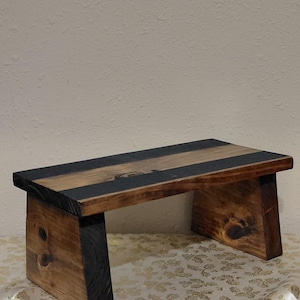 Op de afbeelding: Een kleine, handgemaakte houten kruk. De kruk heeft een rechthoekige bovenkant met een donkere streep en twee stevige poten. Het hout heeft een natuurlijke afwerking met zichtbare knopen en nerven, en de poten zijn zwart gebeitst.