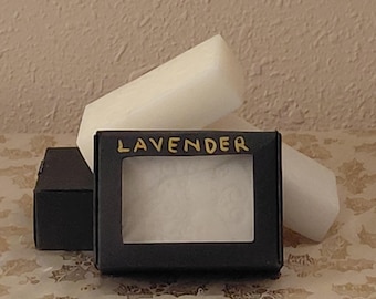 May include: A black box with the word "LAVENDER" in gold lettering. The box has a clear window, revealing a white product. Two white, cylindrical candles are behind the box, and another black box is to the left.