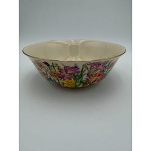 May include: A cream-colored ceramic bowl with a floral design featuring purple, pink, yellow, and orange flowers. The rim of the bowl is trimmed with gold. The bowl is set against a white background.