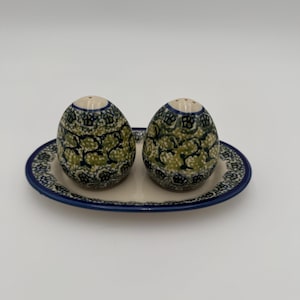 Pode incluir: Um conjunto de saleiro e pimenteiro em cerâmica com dois saleiros em forma de ovo e uma bandeja ovalada a condizer. Os saleiros e a bandeja são decorados com um padrão floral verde e azul sobre um fundo creme, com uma borda azul.