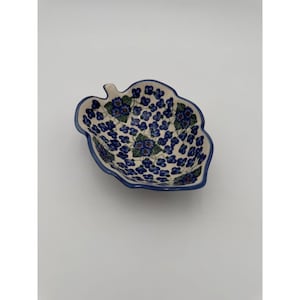 Pode incluir: Um prato de cerâmica em forma de folha com um padrão floral azul. O prato tem uma base creme com uma borda azul e é preenchido com pequenas flores azuis e folhas verdes. O prato tem aproximadamente 15 cm de comprimento e 13 cm de largura.