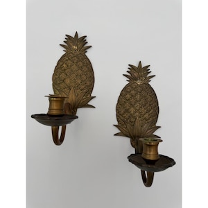 Op de afbeelding: Twee antieke messing ananas wandkandelaars. Elke kandelaar heeft een gedetailleerd ananasontwerp met een kaarsenhouder en een decoratieve bladbasis. De kandelaars zijn gemonteerd op een witte muur, die hun details en warme messing tinten benadrukken.