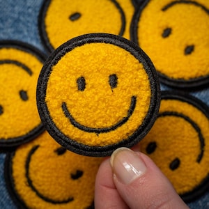May include: A collection of yellow smiley face patches with black embroidered details. The patches have a textured, fuzzy appearance and are round in shape. The patches are being held by a person.