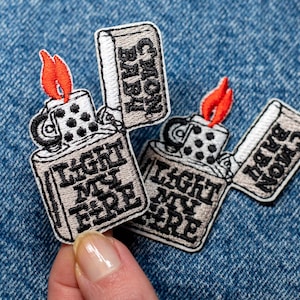 Peut inclure: Écussons brodés représentant un briquet avec une flamme rouge et les textes "LIGHT MY FIRE" et "C'MON BABY" en noir. Les écussons sont posés sur un fond en denim bleu.