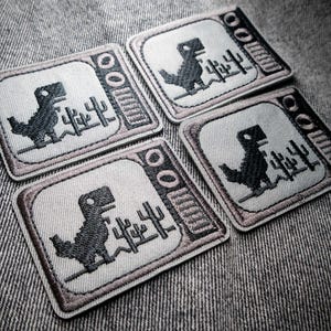 May include: Four embroidered patches featuring a pixelated dinosaur inside a retro television frame. The dinosaur is black, and the frame is outlined in a dark brown. The patches are on a gray fabric background.