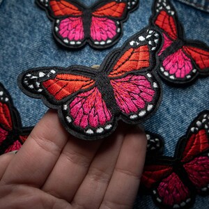 May include: Embroidered butterfly patches in shades of red and pink with black outlines and white accents. The patches are arranged on a denim background, with one patch being held by a hand. These patches are ideal for adding flair to clothing or accessories.