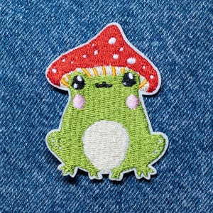 Puede incluir: Parche bordado con una rana caprichosa con un sombrero de hongo. La rana es verde con vientre blanco y mejillas rosas. El sombrero de hongo es rojo con lunares blancos. El parche está sobre un fondo de mezclilla azul.