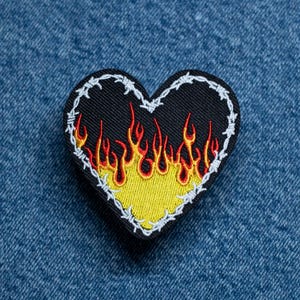 May include: Heart-shaped embroidered patch with a black base, yellow flames, and a barbed wire border. The flames are red and orange. The patch is set against a blue denim fabric background.
