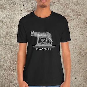May include: Black t-shirt featuring a white graphic of the Capitoline Wolf nursing Romulus and Remus, with the text "ROMA, 753 B.C." The shirt is made of a soft material.