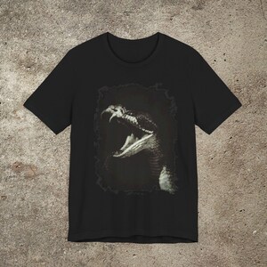 以下が含まれることがあります： 口を開けたヘビのモノクロ画像がプリントされた黒いTシャツ。牙が見えます。ヘビの画像は暗い枠で囲まれています。シャツは柔らかい素材で作られています。