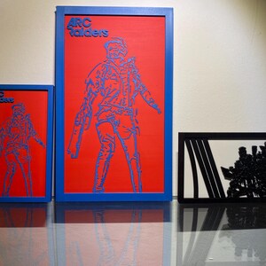 Pode incluir: Três obras de arte emolduradas. Duas apresentam um fundo vermelho com um contorno azul de uma figura, o texto "ARC Raiders" no topo. A terceira é um gráfico a preto e branco de um grupo de figuras.