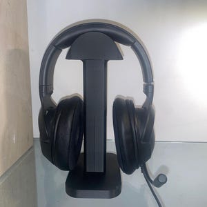 Pode incluir: Fones de ouvido pretos over-ear com microfone embutido em um suporte preto. O suporte é um poste vertical com uma parte superior curva e uma base retangular. Os fones de ouvido têm fones de ouvido grandes e acolchoados.