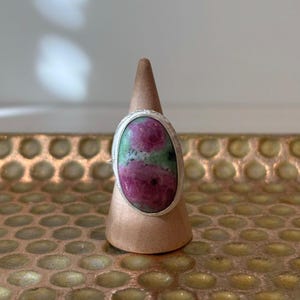 May include: Oval-shaped ring featuring a large, vibrant ruby gemstone with pink and green hues, set in a silver band. The ring is displayed on a copper-colored ring holder, showcasing the jewelry's design and color contrast.